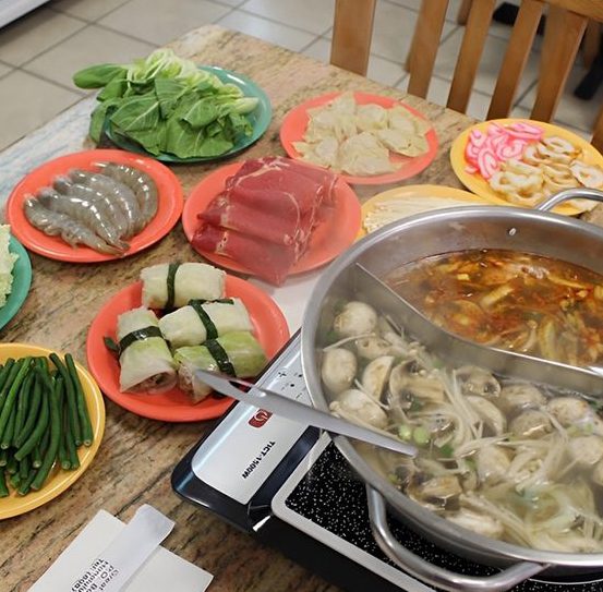 Rectangle-81 A hot pot meal on a table with a bubbling broth in a pot, surrounded by plates of shrimp, beef, greens, rice paper rolls, fish cakes, and beans.