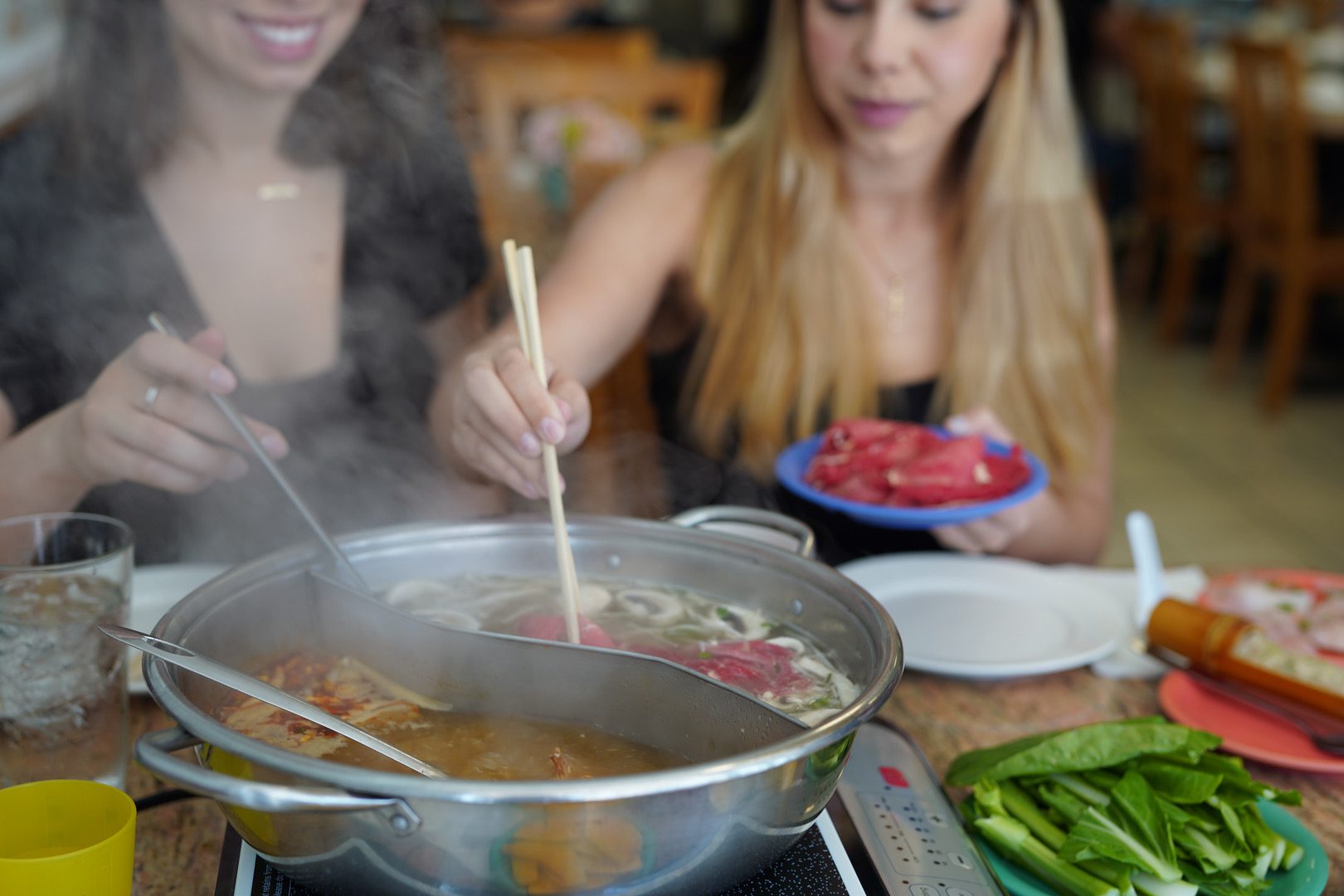 IMG_4587 Two people sit at a table cooking thin slices of meat in a steaming hot pot, surrounded by fresh vegetables and other ingredients.