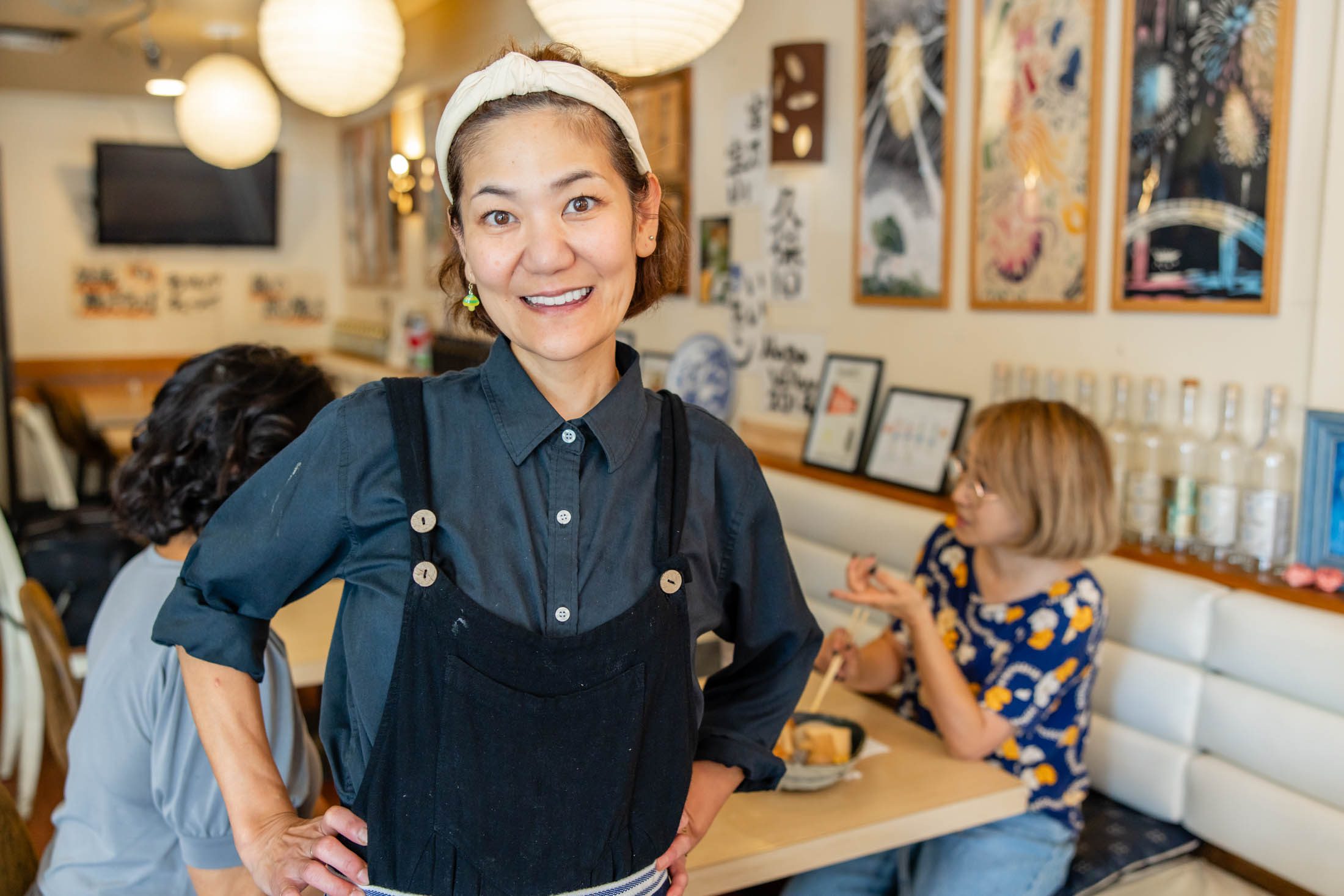 A woman wearing a headband and apron stands smiling in a restaurant, while two people eat at a table in the background.
