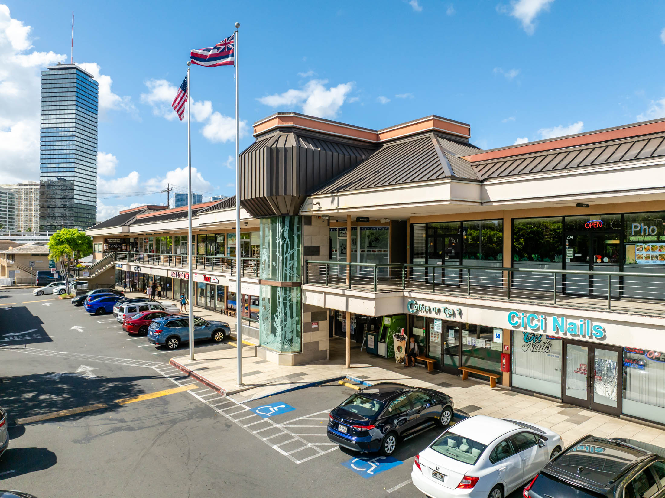 A two-story shopping plaza with parked cars, various storefronts including CICI Nails, and an American flag, with a tall office building in the background under a blue sky.