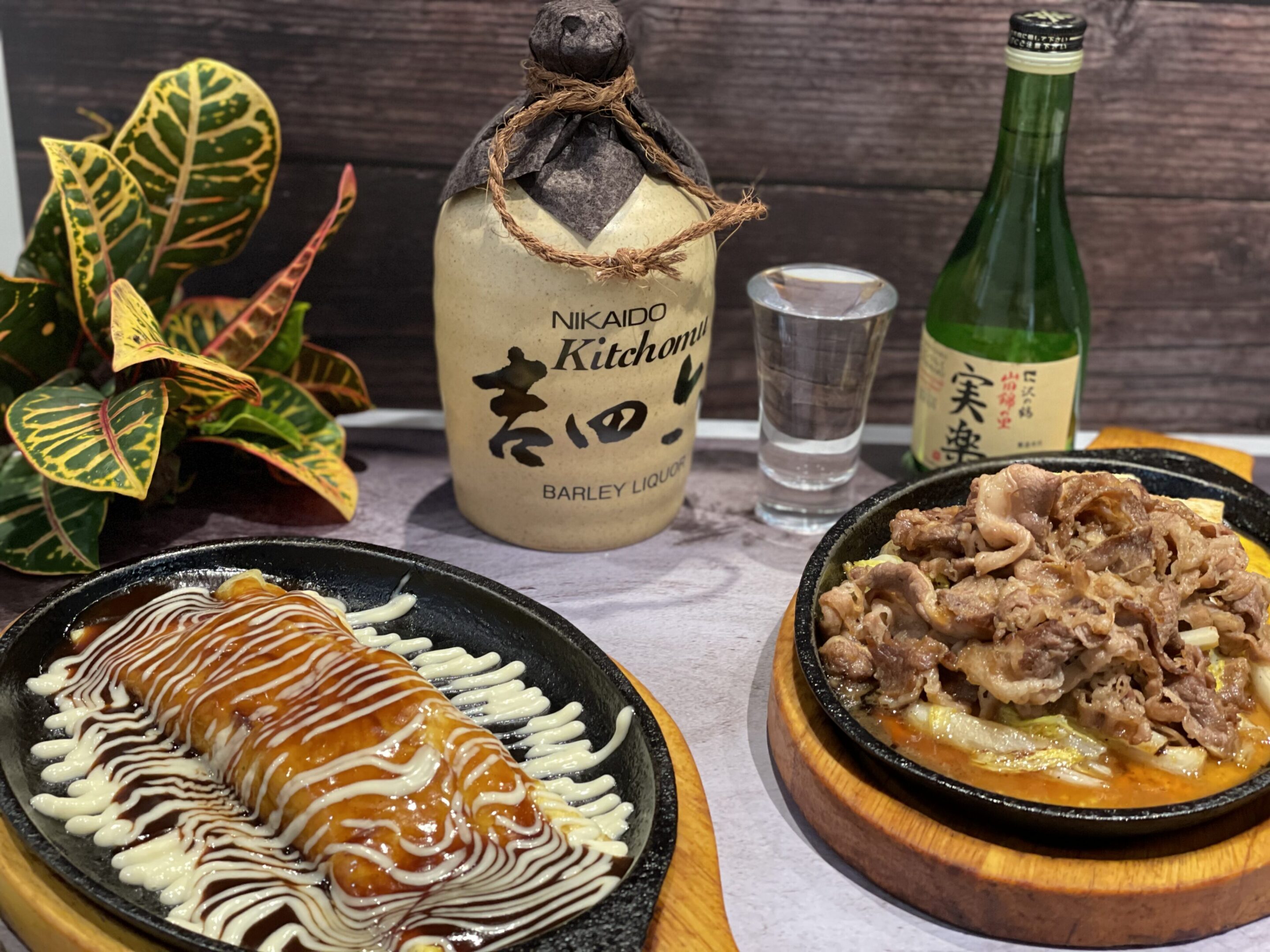 Two sizzling plates of Japanese food with sauces, a bottle of barley liquor, a bottle of sake, a shot glass, and a leafy plant on a wooden table.